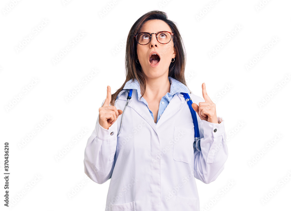 Young beautiful woman wearing doctor stethoscope and glasses amazed and surprised looking up and pointing with fingers and raised arms. Young beautiful woman wearing doctor stethoscope and glasses amazed and surprised looking up and pointing with fingers and raised arms.