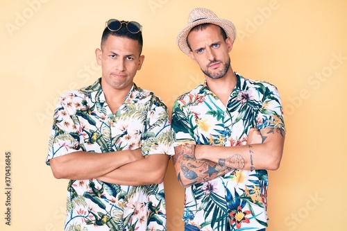 Young gay couple of two men wearing summer hat and hawaiian shirt skeptic and nervous, disapproving expression on face with crossed arms. negative person.