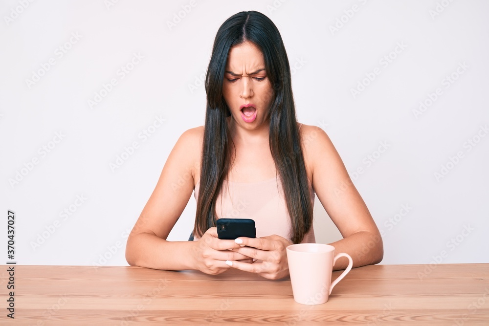 Young caucasian woman sitting at the desk using smartphone drinking coffee scared and amazed with open mouth for surprise, disbelief face Young caucasian woman sitting at the desk using smartphone drinking coffee scared and amazed with open mouth for surprise, disbelief face