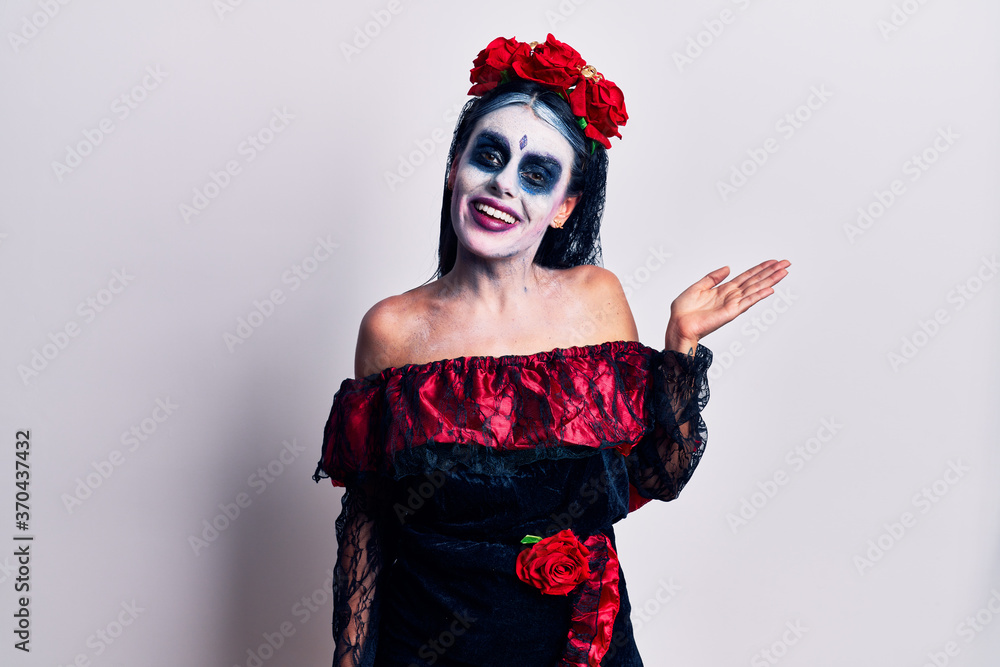 Young woman wearing mexican day of the dead makeup smiling cheerful presenting and pointing with palm of hand looking at the camera. Young woman wearing mexican day of the dead makeup smiling cheerful presenting and pointing with palm of hand looking at the camera.