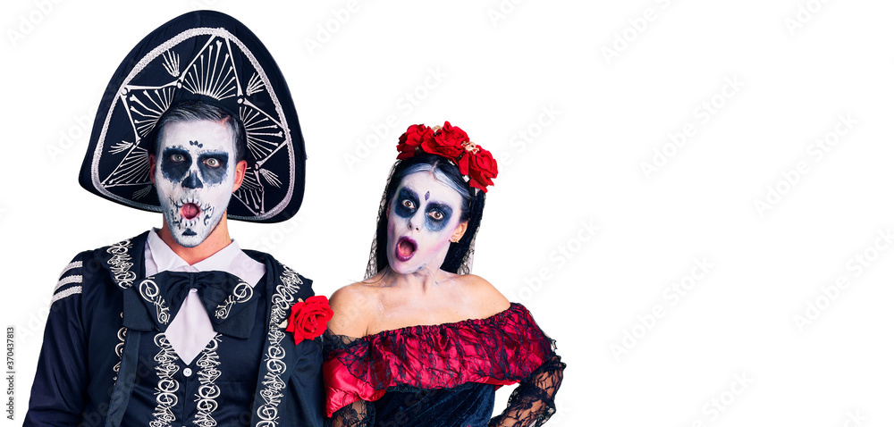 Young couple wearing mexican day of the dead costume over background in shock face, looking skeptical and sarcastic, surprised with open mouth