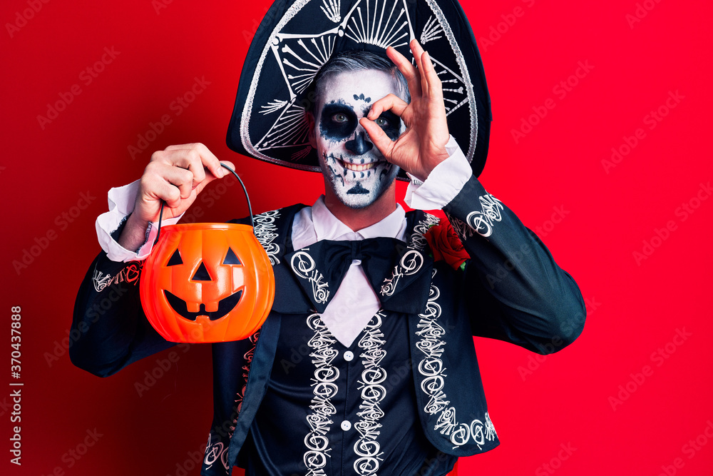 Young man wearing mexican day of the dead costume holding pumpkin smiling happy doing ok sign with hand on eye looking through fingers