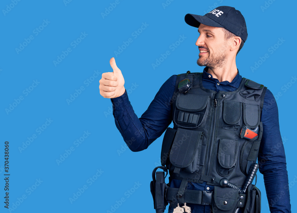 Young handsome man wearing police uniform looking proud, smiling doing thumbs up gesture to the side Young handsome man wearing police uniform looking proud, smiling doing thumbs up gesture to the side