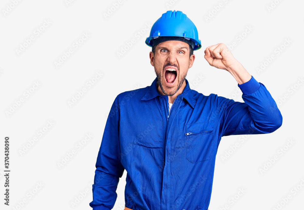 Young handsome man wearing worker uniform and hardhat crazy and mad shouting and yelling with aggressive expression and arms raised. frustration concept. Young handsome man wearing worker uniform and hardhat crazy and mad shouting and yelling with aggressive expression and arms raised. frustration concept.
