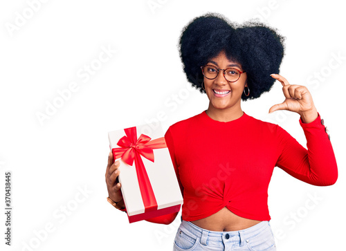 Young african american girl holding gift smiling and confident gesturing with hand doing small size sign with fingers looking and the camera. measure concept.