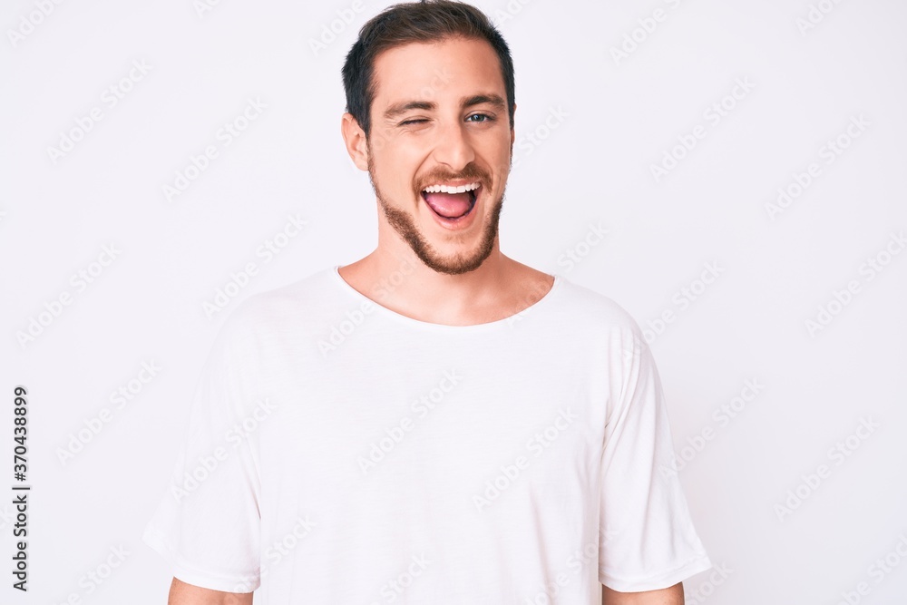 Young handsome man wearing casual white tshirt winking looking at the camera with sexy expression, cheerful and happy face. Young handsome man wearing casual white tshirt winking looking at the camera with sexy expression, cheerful and happy face.