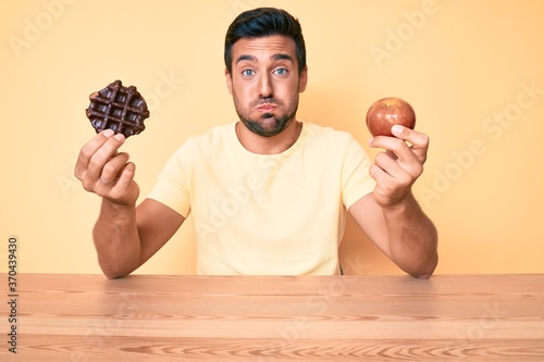 Young hispanic man eating breakfast holding chocolate waflle and apple puffing cheeks with funny face. mouth inflated with air, catching air. photo