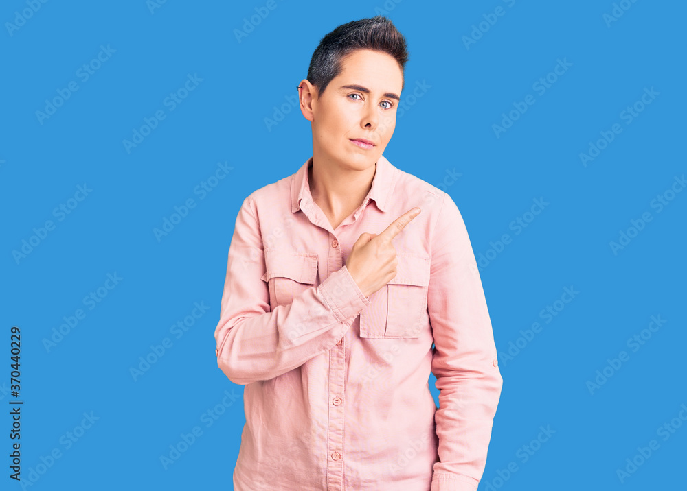 Young woman with short hair wearing casual clothes pointing with hand finger to the side showing advertisement, serious and calm face Young woman with short hair wearing casual clothes pointing with hand finger to the side showing advertisement, serious and calm face