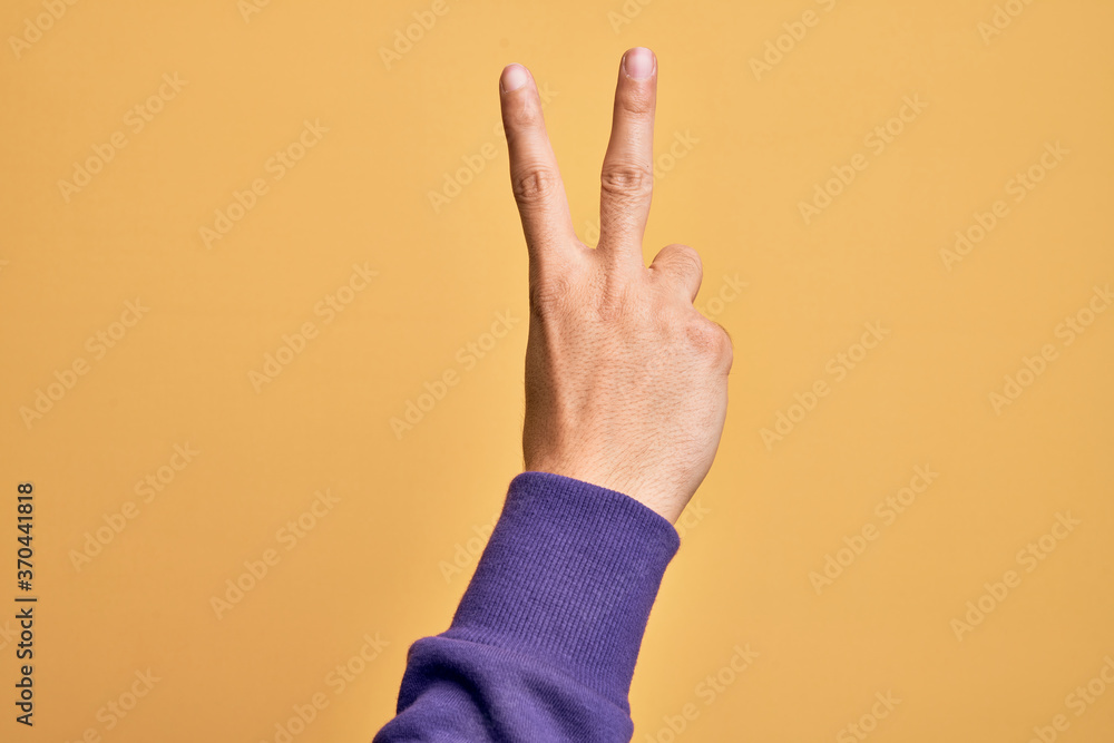 Hand of caucasian young man showing fingers over isolated yellow background counting number 2 showing two fingers, gesturing victory and winner symbol Hand of caucasian young man showing fingers over isolated yellow background counting number 2 showing two fingers, gesturing victory and winner symbol