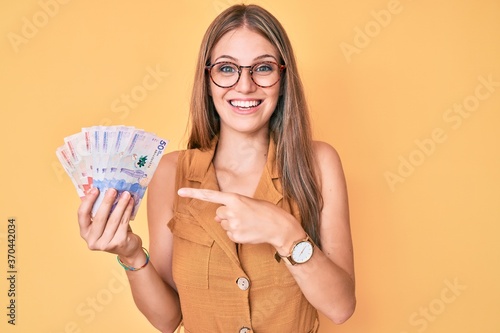 Young blonde girl holding colombian pesos smiling happy pointing with hand and finger photo