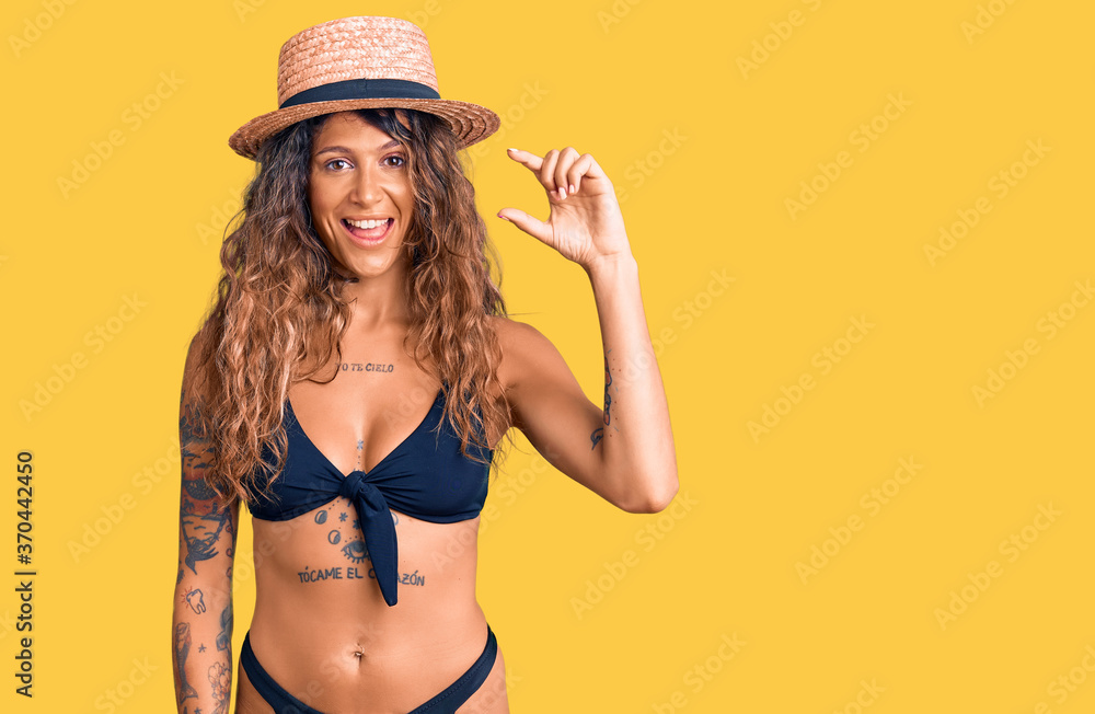 Young hispanic woman with tattoo wearing bikini and summer hat smiling and confident gesturing with hand doing small size sign with fingers looking and the camera. measure concept. Young hispanic woman with tattoo wearing bikini and summer hat smiling and confident gesturing with hand doing small size sign with fingers looking and the camera. measure concept.