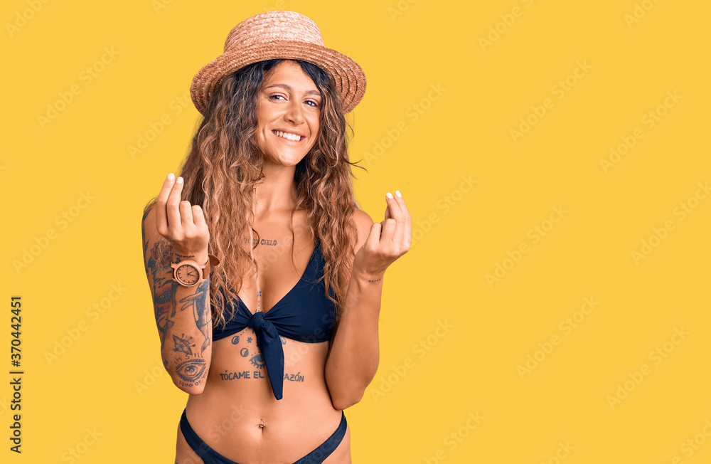 Young hispanic woman with tattoo wearing bikini and summer hat doing money gesture with hands, asking for salary payment, millionaire business
