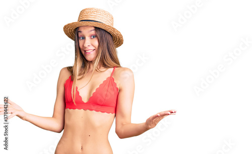 Beautiful brunette young woman wearing bikini clueless and confused expression with arms and hands raised. doubt concept.