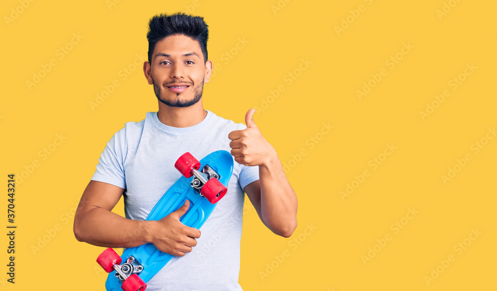 Handsome latin american young man holding skate smiling happy and positive, thumb up doing excellent and approval sign Handsome latin american young man holding skate smiling happy and positive, thumb up doing excellent and approval sign