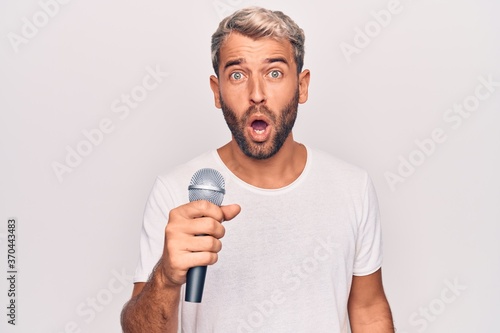 Handsome blond singer man with beard singing song using microphone over white background scared and amazed with open mouth for surprise, disbelief face