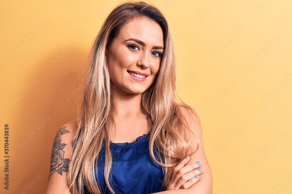 Young beautiful blonde woman wearing casual t-shirt standing over isolated yellow background happy face smiling with crossed arms looking at the camera. Positive person. Young beautiful blonde woman wearing casual t-shirt standing over isolated yellow background happy face smiling with crossed arms looking at the camera. Positive person.