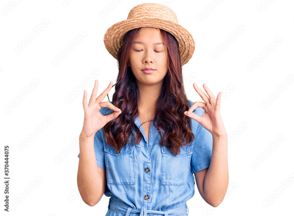 Young beautiful chinese girl wearing summer hat relaxed and smiling with eyes closed doing meditation gesture with fingers. yoga concept. Young beautiful chinese girl wearing summer hat relaxed and smiling with eyes closed doing meditation gesture with fingers. yoga concept.