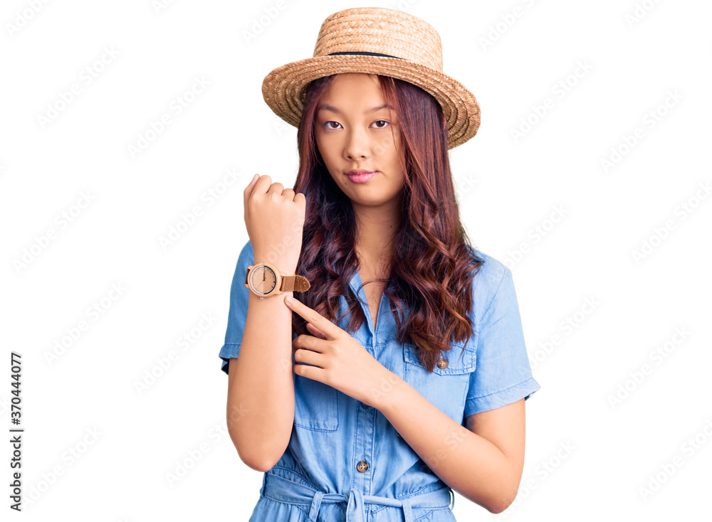 Young beautiful chinese girl wearing summer hat in hurry pointing to watch time, impatience, looking at the camera with relaxed expression Young beautiful chinese girl wearing summer hat in hurry pointing to watch time, impatience, looking at the camera with relaxed expression