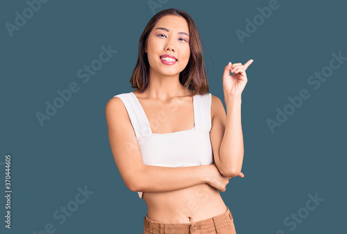 Young beautiful chinese girl wearing casual clothes with a big smile on face, pointing with hand and finger to the side looking at the camera.