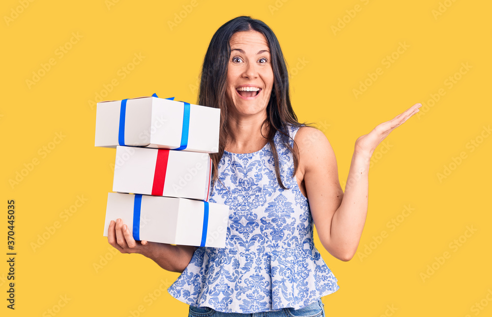 Young beautiful brunette woman holding birthday gift celebrating victory with happy smile and winner expression with raised hands Young beautiful brunette woman holding birthday gift celebrating victory with happy smile and winner expression with raised hands