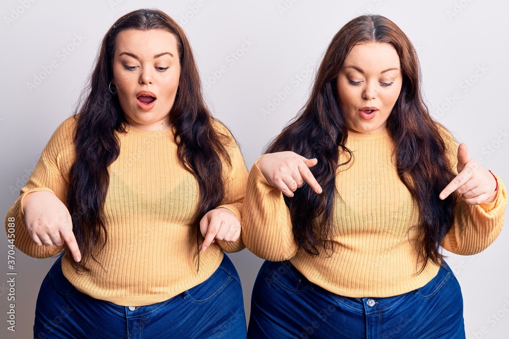 Young plus size twins wearing casual clothes pointing down with fingers showing advertisement, surprised face and open mouth Young plus size twins wearing casual clothes pointing down with fingers showing advertisement, surprised face and open mouth