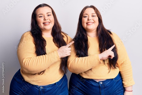 Young plus size twins wearing casual clothes smiling and looking at the camera pointing with two hands and fingers to the side.