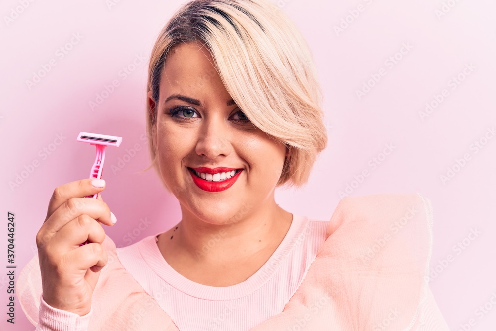 Young beautiful blonde plus size woman holding depilation razor over isolated pink background looking positive and happy standing and smiling with a confident smile showing teeth Young beautiful blonde plus size woman holding depilation razor over isolated pink background looking positive and happy standing and smiling with a confident smile showing teeth