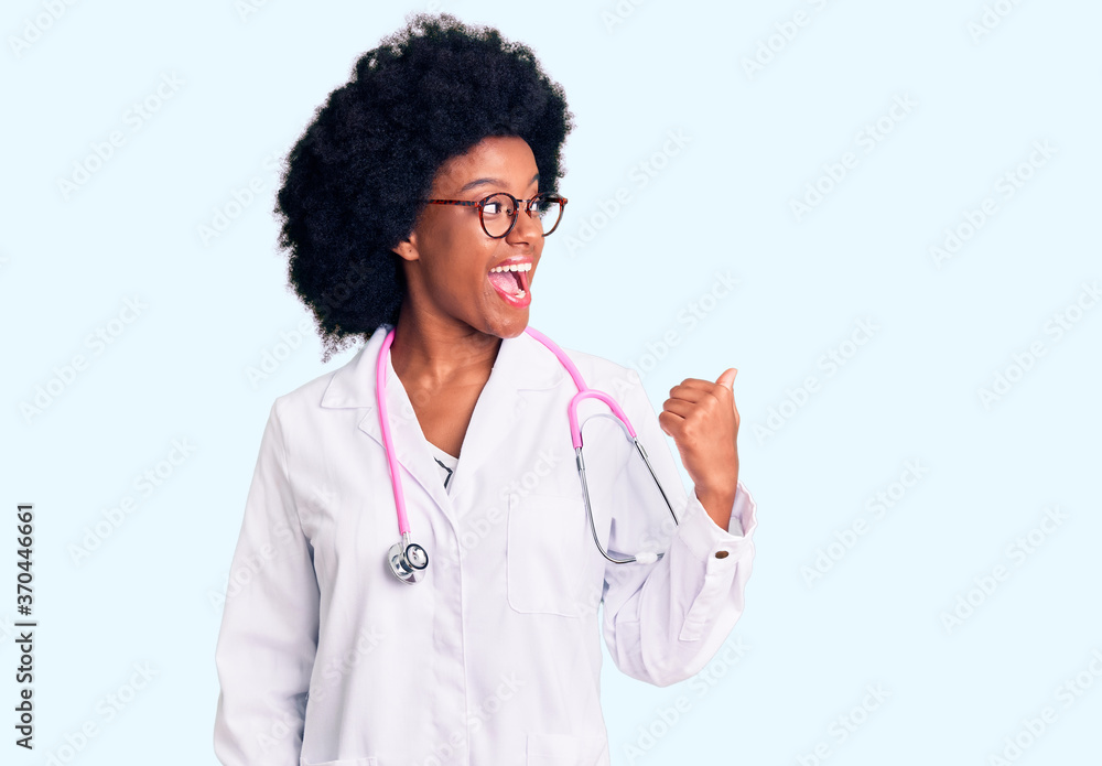 Young african american woman wearing doctor coat and stethoscope smiling with happy face looking and pointing to the side with thumb up. Young african american woman wearing doctor coat and stethoscope smiling with happy face looking and pointing to the side with thumb up.