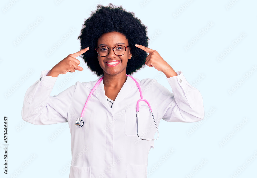 Young african american woman wearing doctor coat and stethoscope smiling pointing to head with both hands finger, great idea or thought, good memory Young african american woman wearing doctor coat and stethoscope smiling pointing to head with both hands finger, great idea or thought, good memory