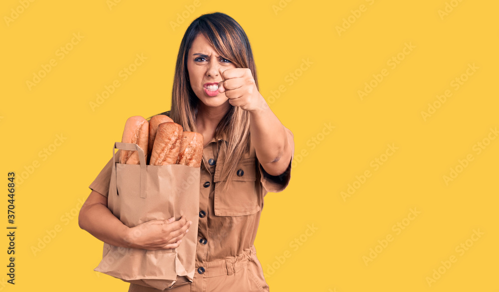 Young beautiful woman holding paper bag with bread annoyed and frustrated shouting with anger, yelling crazy with anger and hand raised Young beautiful woman holding paper bag with bread annoyed and frustrated shouting with anger, yelling crazy with anger and hand raised