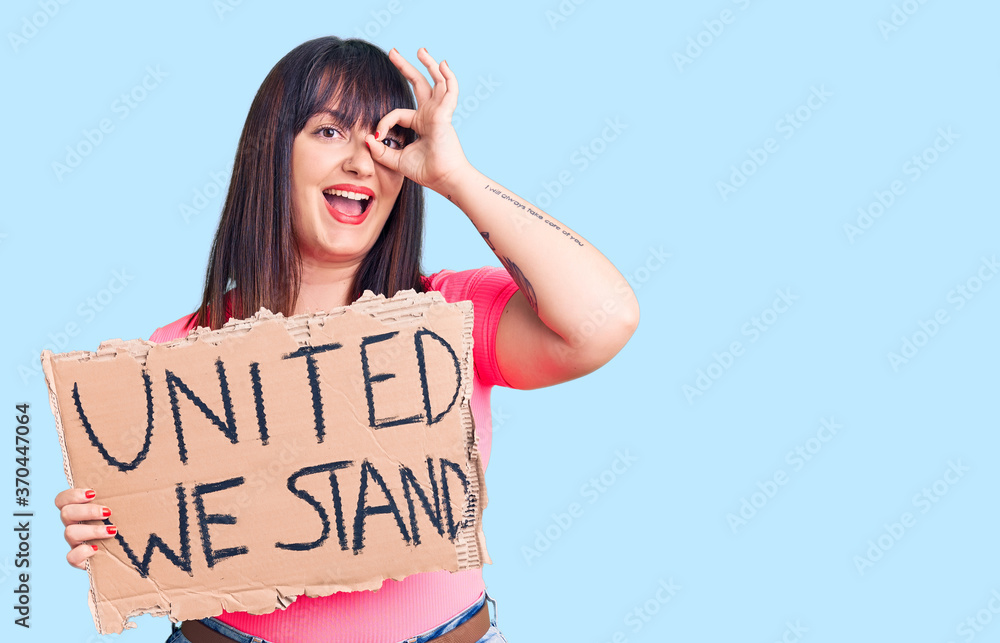 Young plus size woman holding united we stand banner smiling happy doing ok sign with hand on eye looking through fingers Young plus size woman holding united we stand banner smiling happy doing ok sign with hand on eye looking through fingers