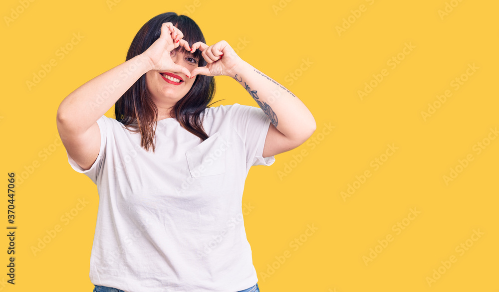 Young plus size woman wearing casual clothes doing heart shape with hand and fingers smiling looking through sign Young plus size woman wearing casual clothes doing heart shape with hand and fingers smiling looking through sign