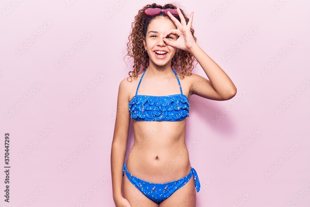 Naklejka premium Beautiful kid girl with curly hair wearing bikini and sunglasses doing ok gesture with hand smiling, eye looking through fingers with happy face.
