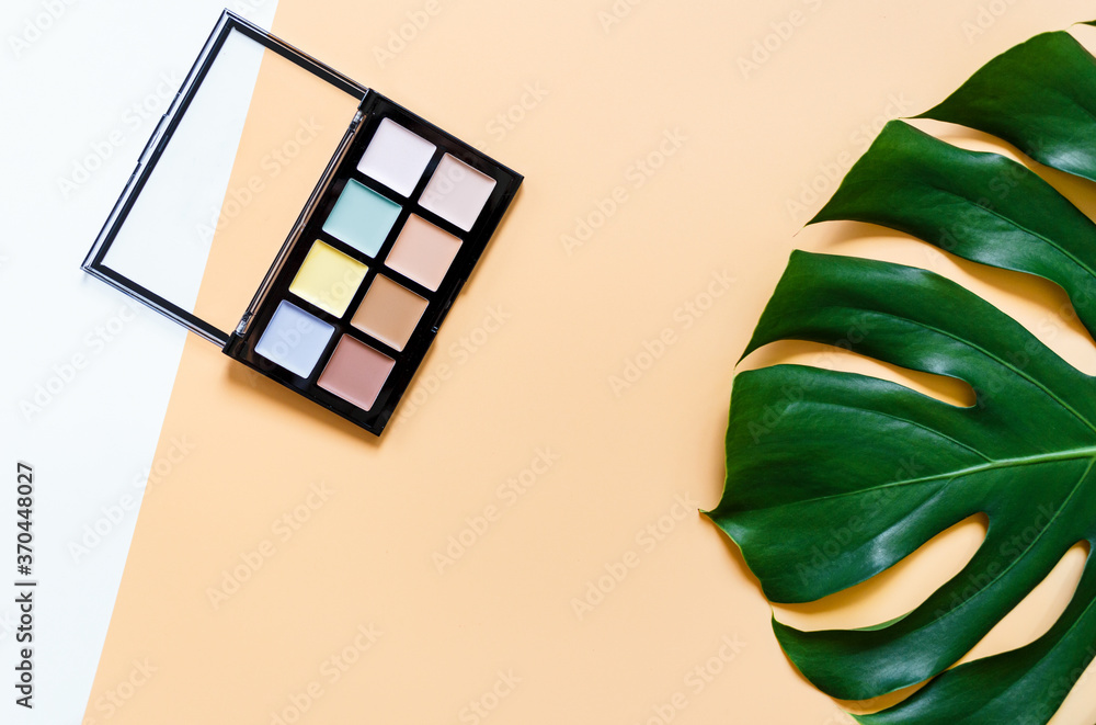 Eye shadow palette on yellow background with big monstera leaf. Beauty, fashion, party and shopping concept. 