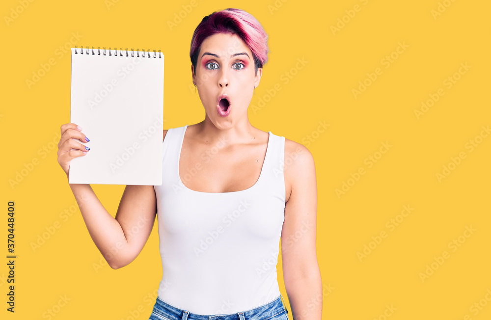 Young beautiful woman with pink hair holding notebook scared and amazed with open mouth for surprise, disbelief face Young beautiful woman with pink hair holding notebook scared and amazed with open mouth for surprise, disbelief face
