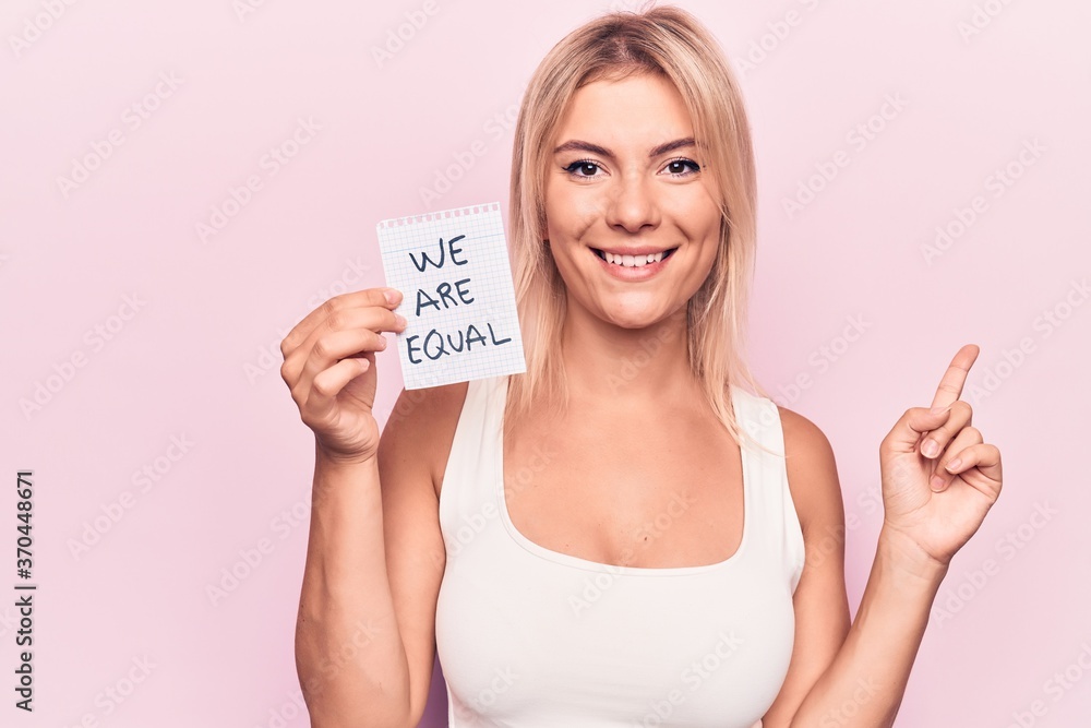 Young beautiful blonde woman asking for equality holding paper with we are equal message smiling happy pointing with hand and finger to the side Young beautiful blonde woman asking for equality holding paper with we are equal message smiling happy pointing with hand and finger to the side