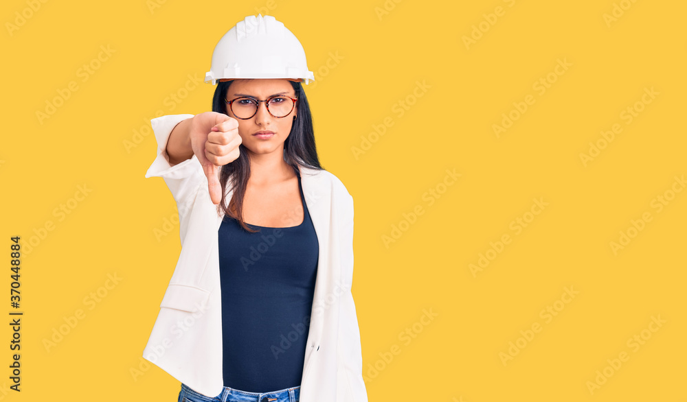 Young beautiful latin girl wearing architect hardhat and glasses looking unhappy and angry showing rejection and negative with thumbs down gesture. bad expression.