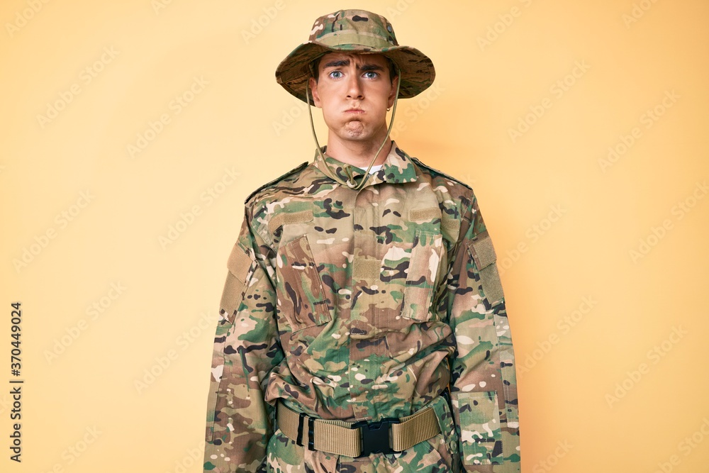 Young handsome man wearing camouflage army uniform puffing cheeks with funny face. mouth inflated with air, crazy expression. Young handsome man wearing camouflage army uniform puffing cheeks with funny face. mouth inflated with air, crazy expression.