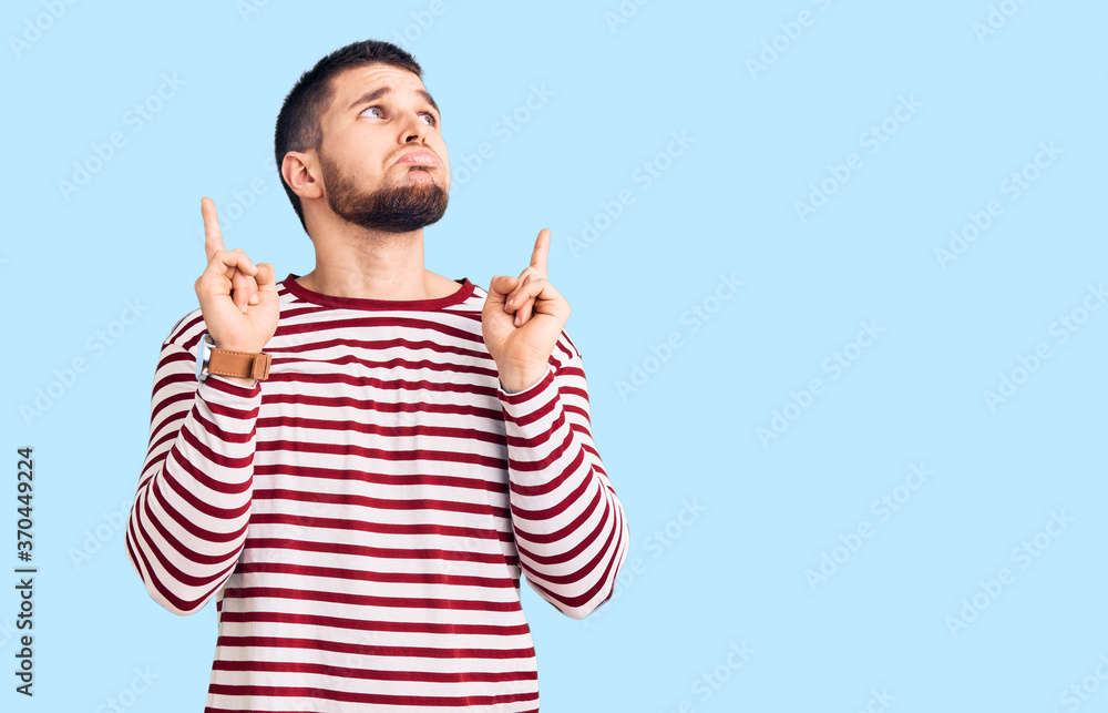 Young handsome man wearing striped sweater pointing up looking sad and upset, indicating direction with fingers, unhappy and depressed. Young handsome man wearing striped sweater pointing up looking sad and upset, indicating direction with fingers, unhappy and depressed.