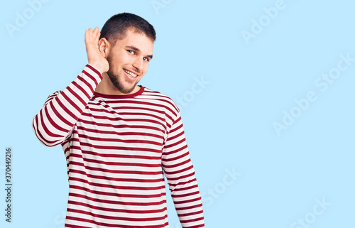 Young handsome man wearing striped sweater smiling with hand over ear listening an hearing to rumor or gossip. deafness concept.