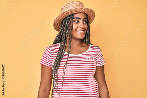 Young african american woman with braids wearing summer hat looking away to side with smile on face, natural expression. laughing confident.