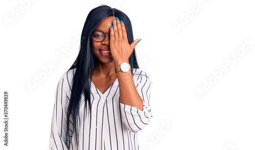 Young african american woman wearing casual clothes and glasses covering one eye with hand, confident smile on face and surprise emotion.