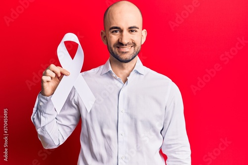 Young handsome bald man holding white cancer ribbon looking positive and happy standing and smiling with a confident smile showing teeth photo