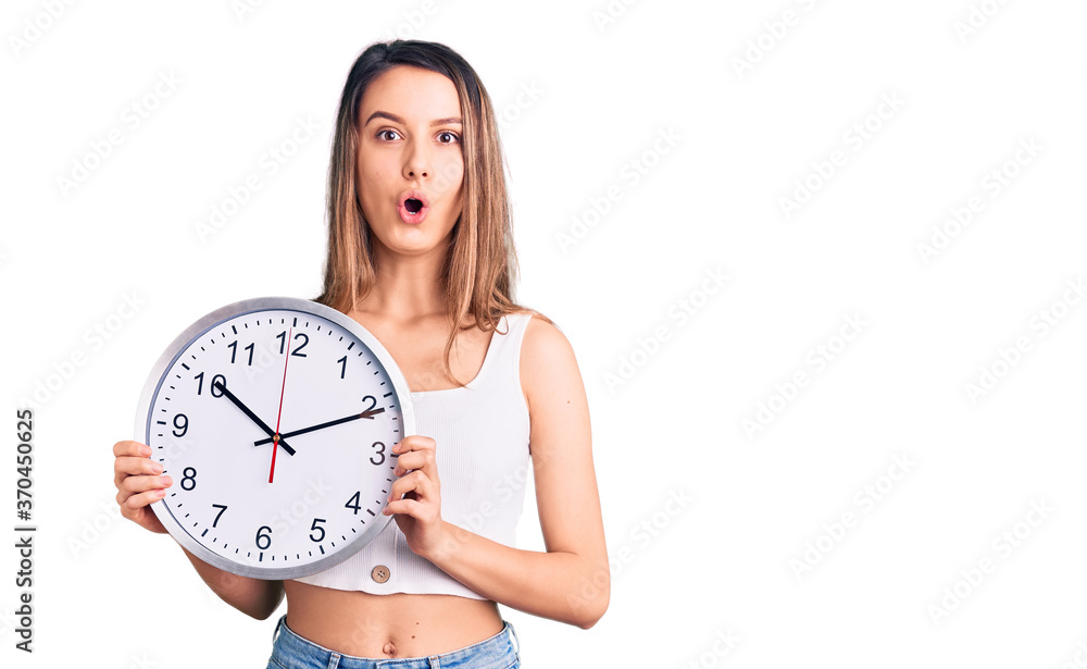 Young beautiful girl holding big clock scared and amazed with open mouth for surprise, disbelief face Young beautiful girl holding big clock scared and amazed with open mouth for surprise, disbelief face