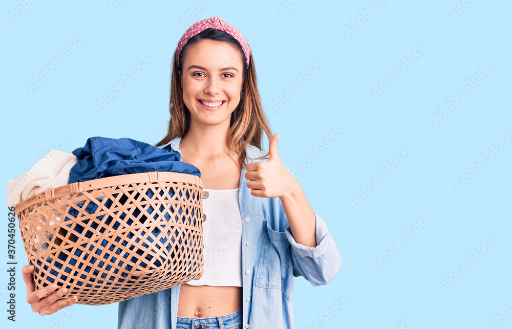 Young beautiful girl holding laundry basket smiling happy and positive, thumb up doing excellent and approval sign