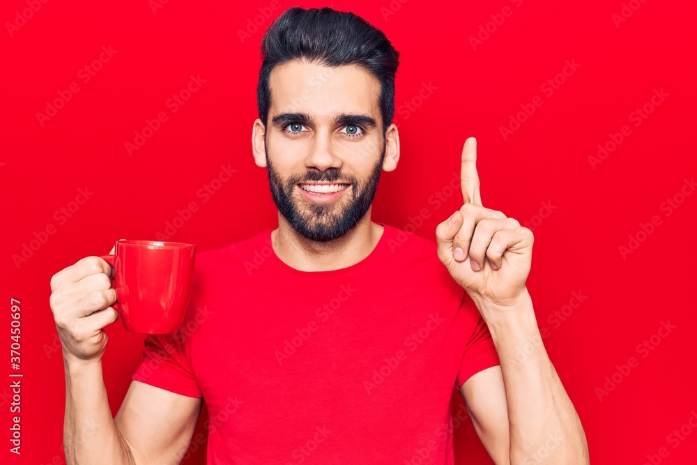 Young handsome man with beard drinking mug of coffee smiling with an idea or question pointing finger with happy face, number one Young handsome man with beard drinking mug of coffee smiling with an idea or question pointing finger with happy face, number one