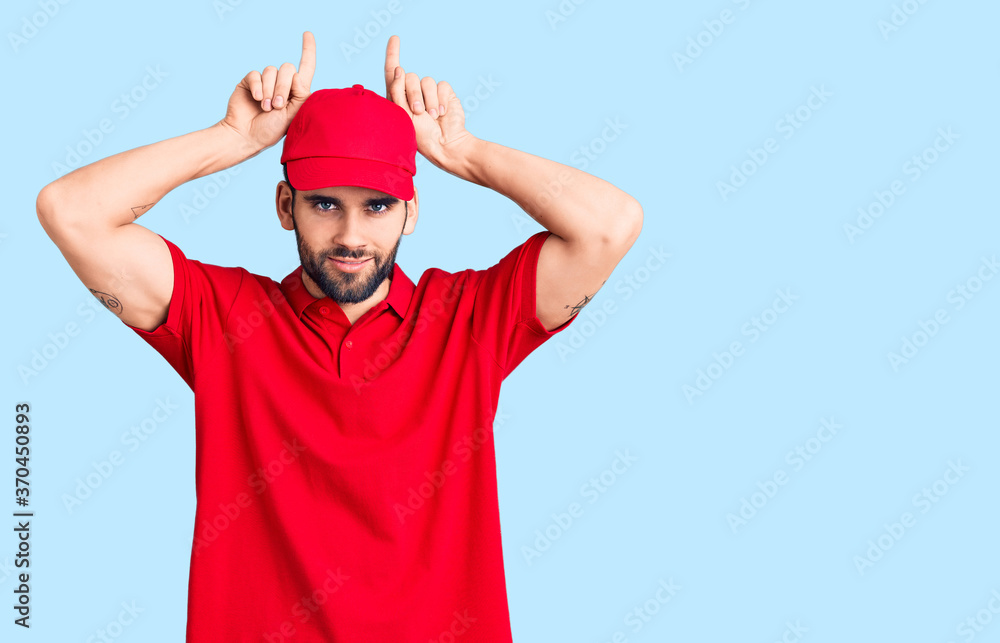 Naklejka premium Young handsome man with beard wearing delivery uniform doing funny gesture with finger over head as bull horns