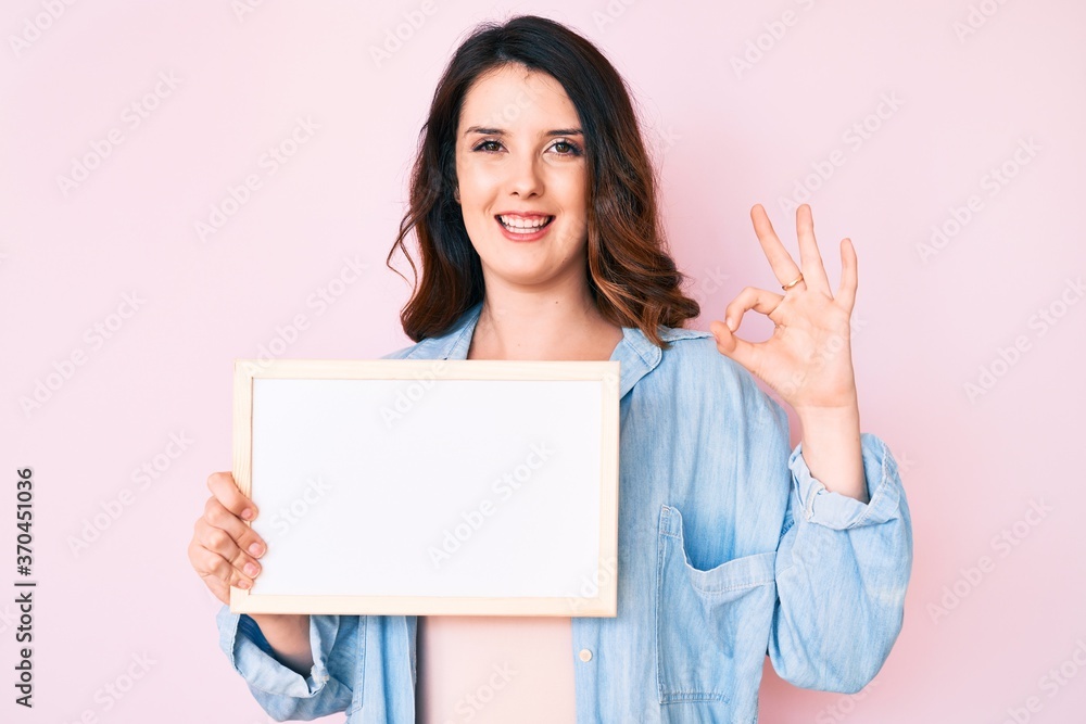 Young beautiful brunette woman holding blank empty banner doing ok sign with fingers, smiling friendly gesturing excellent symbol Young beautiful brunette woman holding blank empty banner doing ok sign with fingers, smiling friendly gesturing excellent symbol