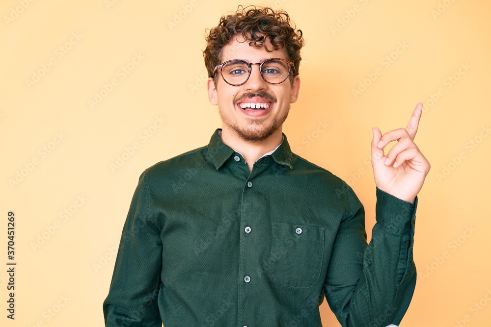 Young caucasian man with curly hair wearing casual clothes and glasses with a big smile on face, pointing with hand and finger to the side looking at the camera. Young caucasian man with curly hair wearing casual clothes and glasses with a big smile on face, pointing with hand and finger to the side looking at the camera.