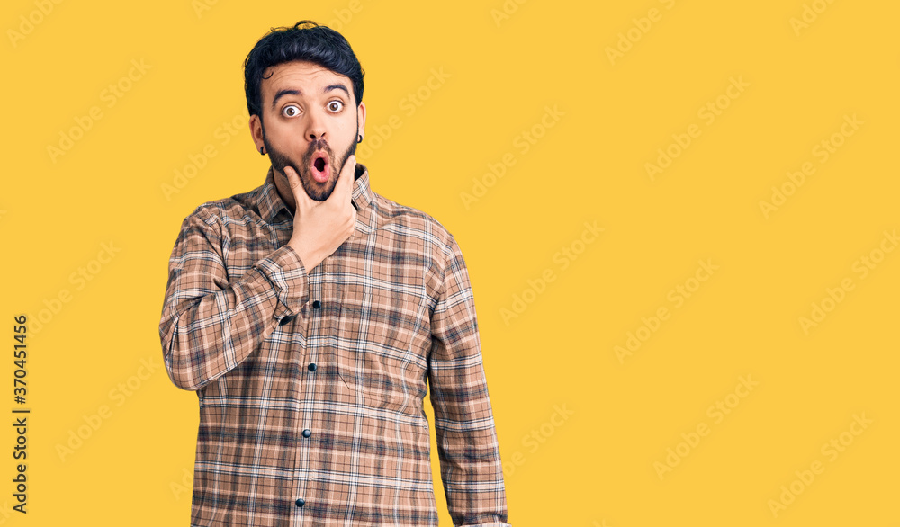 Young hispanic man wearing casual clothes looking fascinated with disbelief, surprise and amazed expression with hands on chin Young hispanic man wearing casual clothes looking fascinated with disbelief, surprise and amazed expression with hands on chin